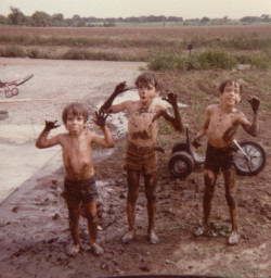 three brothers playing in the mud. There is a driveway to the left of them, a big wheel behind them, a field in the background
