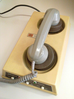 a large rectangular phone modem. The modem is beige in color and then phone placed into it is grey. There is a black electric cord extruding from the top left and a coiled phone cord coming out of the microphone side of the headset protruding toward the left off screen. The apparatus sits on what appears to be a white desk or counter