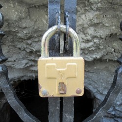 Photo of a gold color padlock with silver u-bolt. It is slightly rusty on the bottom. The lock has changed the opening of a wrought iron gate in front of what appears to be a rock wall with an opening in it.