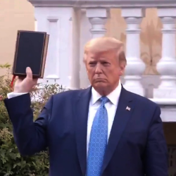 Donald Trump holding up a Bible with his right arm and hand. He is wearing a dark blue suit, light blue tie, and on his left chest a pin of the American flag. In the background is St John's Church in Washington DC.